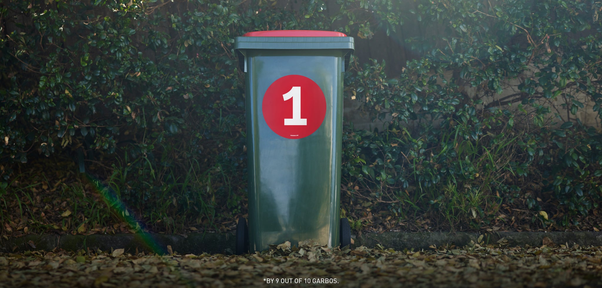  Red wheelie bin in leaflitter with green hedge behind with a red number 1 Suburbin sticker.